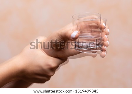 A closeup view on the hands of a person trying to hold a glass of water steady, shaking hands symptomatic of a central nervous and motor system disease such as Parkinson's