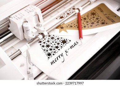 closeup view on machine head of plotting machine that cuts christmas sticker with lettering from black adhesive vinyl. DIY concept with tools and additional sticker. warm light mood. selective focus