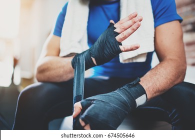 Closeup View On Male Hands Of Young Athlete Tying Black Boxing Bandages.Boxer Man Relaxing After Hard Kickboxing Training Session In Gym. Blurred Background. Horizontal