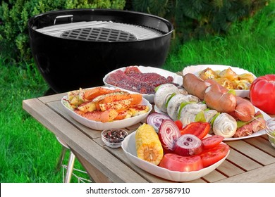 Close-up View On Wood Picnic Table  With Different Cookout Food For Summer BBQ Family Party On The Backyard And Empty Barbecue Grill Appliance On The Green Grass And Plants Background