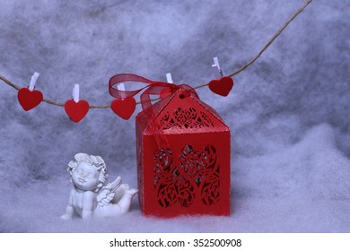 Closeup view of one beautiful cupid angel decorative figurine near red paper greeting valentine box and hanging clothes-peg in shape of heart with white wadding decorating snow, horizontal picture - Powered by Shutterstock