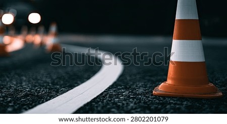 A close-up view of an orange and white traffic cone on a dark asphalt road at night, with a white line curving into the distance.