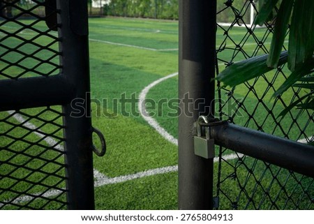 Close-up view of a padlock attached to the black fence gate of a mini soccer field. The field features green artificial turf and is surrounded by lush trees and plants, representing security, restricted access, and outdoor sports facility concepts.