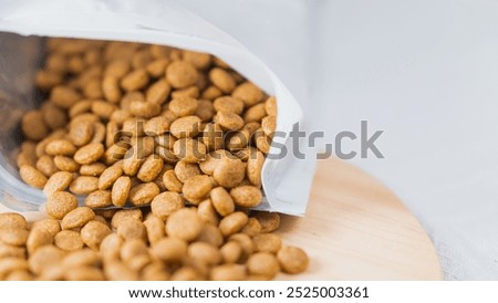 A close-up view of a partially opened bag of pet food, with kibble spilling out onto a wooden surface. The kibble is small, round, and brown, suggesting it is for dogs or