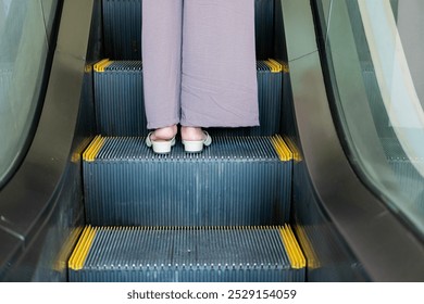 A close-up view of a person's feet on an escalator. The individual is wearing light-colored sandals and loose-fitting, gray pants. The escalator steps are visible with ye - Powered by Shutterstock