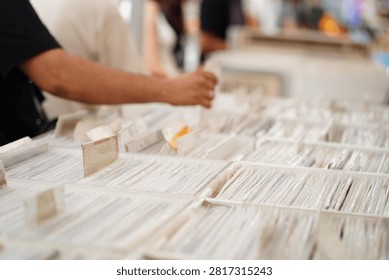 Close-up view of a person's hand browsing through a collection of vinyl records in a bright indoor music store. The hand gently pulls a record sleeve from a crowded bin, with a gold wedding band visible on the finger. Shallow depth of field creates a soft bokeh background, highlighting the nostalgic experience of discovering music, vintage records, and physical media. Clean composition with natural lighting and detailed textures of vinyl covers.