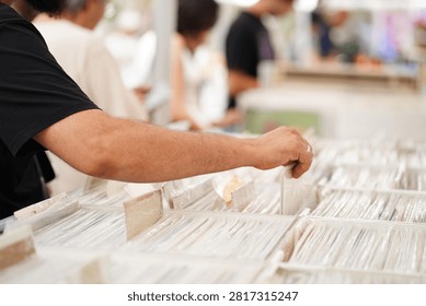 Close-up view of a person's hand browsing through a collection of vinyl records in a bright indoor music store. The hand gently pulls a record sleeve from a crowded bin, with a gold wedding band visible on the finger. Shallow depth of field creates a soft bokeh background, highlighting the nostalgic experience of discovering music, vintage records, and physical media. Clean composition with natural lighting and detailed textures of vinyl covers.