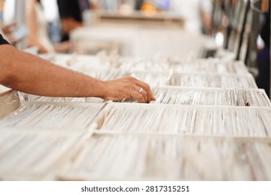 Close-up view of a person's hand browsing through a collection of vinyl records in a bright indoor music store. The hand gently pulls a record sleeve from a crowded bin, with a gold wedding band visible on the finger. Shallow depth of field creates a soft bokeh background, highlighting the nostalgic experience of discovering music, vintage records, and physical media. Clean composition with natural lighting and detailed textures of vinyl covers.