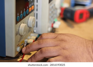 A close-up view of a person's hand operating a benchtop DC power supply unit with control knobs, buttons, and wire terminals.