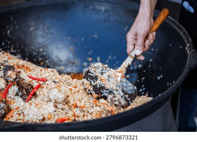Close-up view of a persons hand using a wooden-handled ladle to stir a large cauldron filled with cooked rice mixed with pieces of braised meat, red chili peppers, carrots,