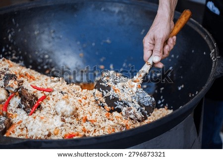 Close-up view of a persons hand using a wooden-handled ladle to stir a large cauldron filled with cooked rice mixed with pieces of braised meat, red chili peppers, carrots,