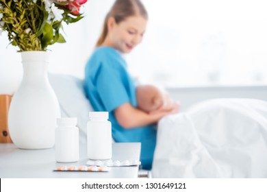 Close-up View Of Pills, Flowers In Vase And Young Mother Breastfeeding Newborn Baby In Hospital Room