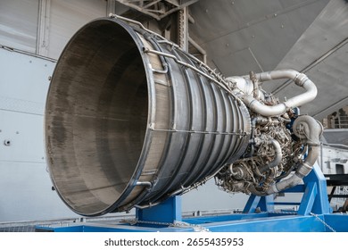 A close-up view of a powerful rocket engine with visible nozzle, pipes, and turbopump components. The engine is mounted on a blue metal frame for exhibition purposes.
