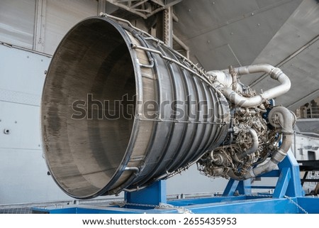 A close-up view of a powerful rocket engine with visible nozzle, pipes, and turbopump components. The engine is mounted on a blue metal frame for exhibition purposes.