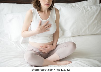 Close-up View Of Pregnant Young Woman Doing Yoga Exercise. Female Model Sitting On The Bed Cross-legged In Easy Pose And Using Breathing Techniques With Hands On Chest And Belly