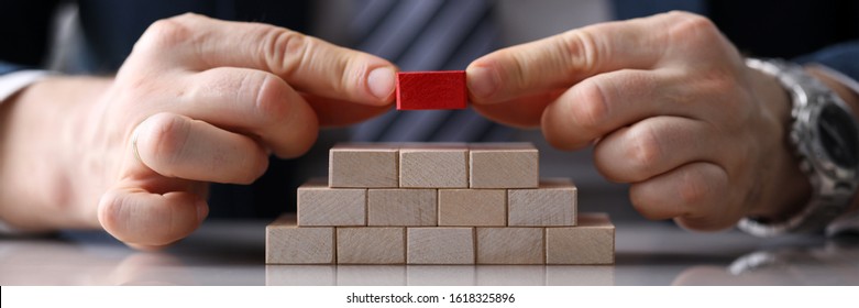 Close-up view of presentable man putting unique red block on staircase of other identical bricks. Pyramid of wooden cubes on desktop. Authority and uniqueness concept