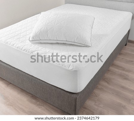 Close-up View of Pristine White Bed, Geometric Quilting on Mattress and Pillow, Contrasting with Soft Gray Upholstered Bed Frame, Comfort, Set Against a Warm Wooden Floor Background. white walls.