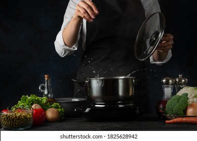 Close-up view of the professional chef in black apron salting water before cooking soup on dark blue background. Backstage of preparing healthy meal. Food concept. Frozen motion.