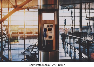 Close-up view of push-button payphone in opened transparent phone booth located in modern airport terminal or contemporary shopping mall interior, or railway depot station with waiting hall behind