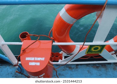 A close-up view of the railing and deck of a ship, bright orange lifebuoy secured by the ropes, safety equipment, with ocean in the background, turquoise water - Powered by Shutterstock