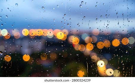 Close-up view of raindrops on a window pane, with blurred city lights creating a bokeh effect in the background during twilight.