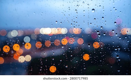 Close-up view of raindrops on a window pane, with blurred city lights creating a bokeh effect in the background.