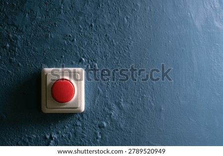 A close-up view of a red emergency button on a rough, textured blue wall, suggesting urgency or a call to action.