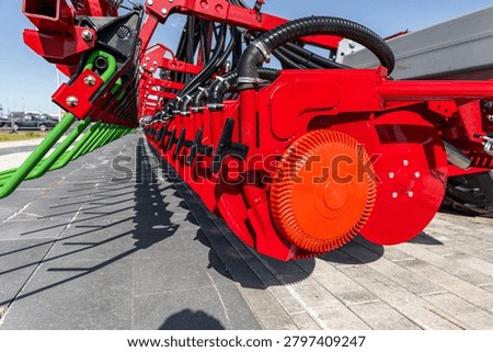 Close-up view of a red seeder machine parked on a paved farmyard, showing metal discs, coulters, gear housing, and hydraulic lines. Bright painted steel
