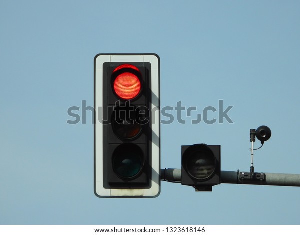 Closeup view of red traffic lights with camera\
tracking and controlling the flow of cars mounted on a grey\
metallic pole above the road, Berlin, Germany. Clear bright blue\
sky in the background