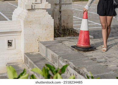 A close-up view of a red and white traffic cone placed on a cobblestone surface, with a blurred background of plants and a parking area marked with white lines. - Powered by Shutterstock