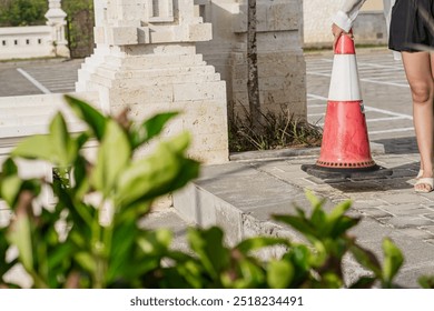 A close-up view of a red and white traffic cone placed on a cobblestone surface, with a blurred background of plants and a parking area marked with white lines. - Powered by Shutterstock