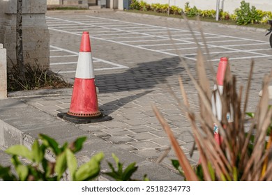 A close-up view of a red and white traffic cone placed on a cobblestone surface, with a blurred background of plants and a parking area marked with white lines. - Powered by Shutterstock