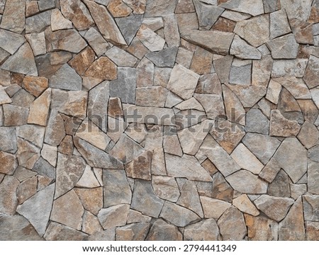 A close-up view of a rough wall built from irregularly shaped stones in a palette of earthy grays, beiges, tans, and browns. The stones create a textured