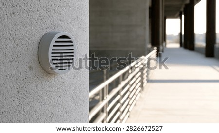 A close-up view of a round, grey ventilation grille attached to a rough concrete wall, with a modern walkway in the background.