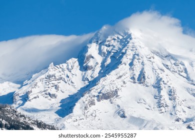 A close-up view of the rugged, snow-covered peaks of Mount Rainier, partially veiled by a delicate layer of clouds under a bright blue sky, capturing the mountain's majesty and serene atmosphere. - Powered by Shutterstock