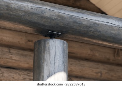 Close-up view of a rustic log cabin timber post supporting a horizontal beam, highlighting natural wood grain, joinery details and a metal plate fastener in warm, weathered architectural texture