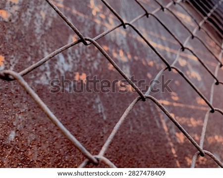 Close-up view of a rusty chain link fence over a weathered metal surface with faded orange markings, creating a rugged industrial texture.