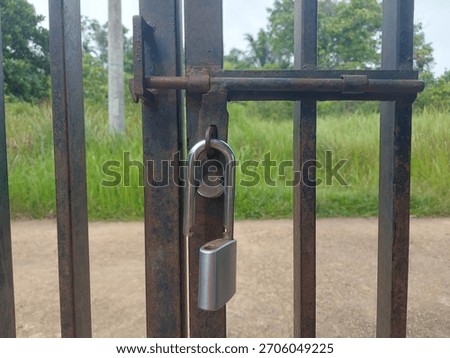 A close-up view of a rusty iron gate secured with a padlock in an outdoor rural area. The worn texture and industrial feel create a strong visual for security, restriction, abandonment, or rustic them