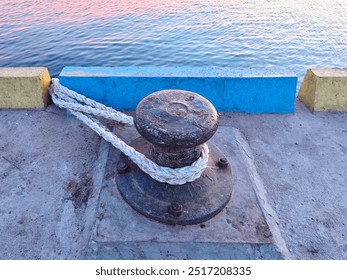 A close-up view of a rusty metal mooring bollard tied with a thick white rope at a concrete dock, with blue and yellow barriers and calm water in the background - Powered by Shutterstock