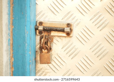 A close-up view of a rusty padlock securing a latch on a metal door featuring a diamond plate texture, with a blue-painted wooden frame visible on the left side.
