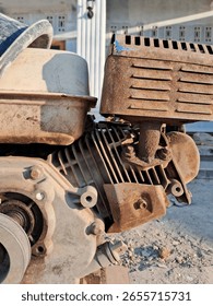 A close-up view of a rusty, weathered engine, likely for machinery or a vehicle, showcasing its metallic texture and components. The background appears to be an outdoor setting with a blurred building