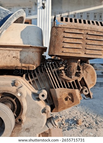 A close-up view of a rusty, weathered engine, likely for machinery or a vehicle, showcasing its metallic texture and components. The background appears to be an outdoor setting with a blurred building