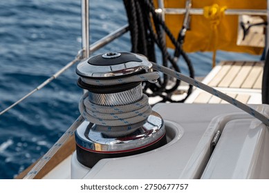 A close-up view of a sailing winch with a rope wrapped around the metallic drum on the deck of a yacht. The image shows detailed nautical equipment with elements of the boat and water visible in the background. It highlights marine hardware, sailing mechanics, and the technical aspects of operating a sailboat.