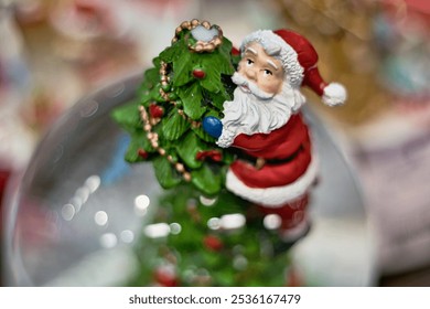 A close-up view of a Santa Claus figurine inside a glass sphere, holding a miniature Christmas tree decorated with gold and red ornaments. - Powered by Shutterstock