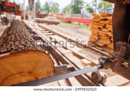 A close-up view of a sawmill with a large log being cut by a band saw. The saw blade is in focus, showing the cutting mechanism, while the log and sawmill environment are blurred in the background.