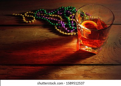 Close-up View Of Sazerac Cocktail With A Lemon Peel In Glass On Wooden Table With Side Lighting And Warm Tones. Mardi Gras Beads In Green, Gold And Purple Nearby.