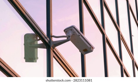 Close-up view of a security camera attached to a modern glass building. The shiny surface reflects orange and pink hues from the sunset sky, showcasing urban architecture.