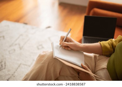 Closeup view of senior woman's hands writing in notebook while sitting on couch beside her laptop, cropped image of elderly lady taking notes to notepad, having moment of creativity or work - Powered by Shutterstock