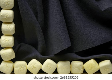 A close-up view showcases a stack of soft, pale yellow marshmallows on the left and a row of similar marshmallows along the bottom edge, set against a draped, textured black fabric.