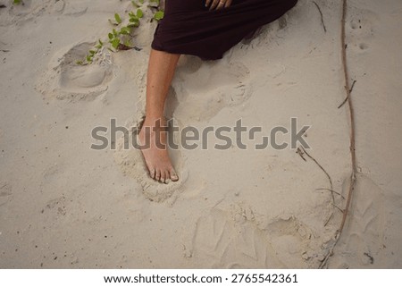 A close-up view shows a person's bare foot pressed into the fine grains of sand, with a small green vine and a piece of driftwood nearby